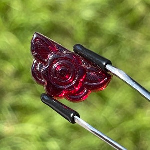 May include: A deep red, rose-shaped gemstone is held by a pair of tweezers. The gemstone has intricate details, including petals and leaves. The tweezers have black rubber tips. The background is a blurred green.