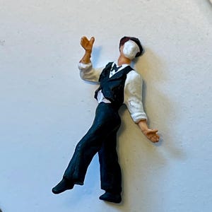 May include: Miniature figurine of a man in a formal outfit, including a white shirt, black vest, and trousers. The figure has a white mask covering the face and is posed with arms outstretched, as if dancing or performing.