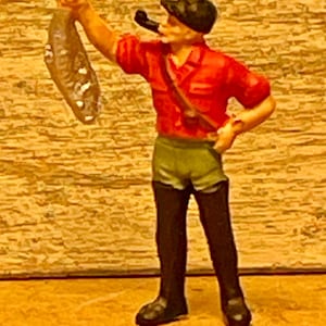 May include: A miniature figurine of a fisherman in a red shirt, green shorts, and black boots, holding up a fish. He wears a black cap and smokes a pipe. The figure is set against a textured, light brown background.