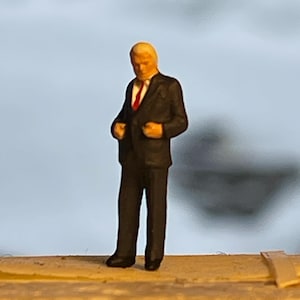 May include: A miniature figurine of a person in a black suit with a red tie. The figure has blonde hair and is standing with hands near the jacket pockets. The background is a blurred light blue.