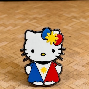 K&ouml;nnte beinhalten: Ein wei&szlig;er Emaille-Pin mit einer stilisierten Katze mit schwarzen Schnurrhaaren und einer Schleife. Die Schleife ist blau, gelb und rot. Das Outfit der Katze ist rot und blau mit einem gelben Stern in der Mitte. Der Pin hat einen silbernen Rand.