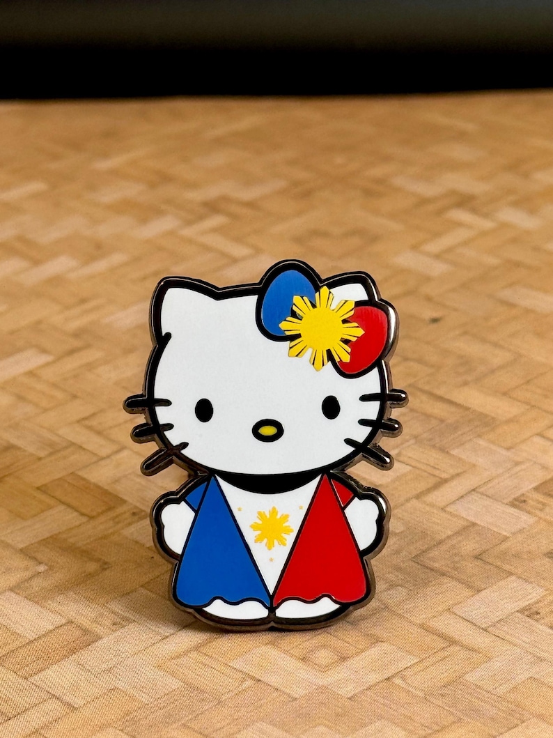 K&ouml;nnte beinhalten: Ein wei&szlig;er Emaille-Pin mit einer stilisierten Katze mit schwarzen Schnurrhaaren und einer Schleife. Die Schleife ist blau, gelb und rot. Das Outfit der Katze ist rot und blau mit einem gelben Stern in der Mitte. Der Pin hat einen silbernen Rand.