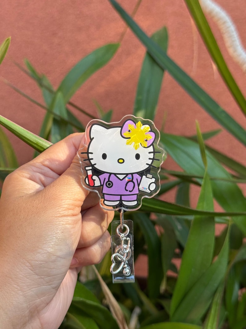 May include: A clear badge reel featuring a cartoon character in a purple medical uniform with a stethoscope. The character has a yellow bow and is holding medical supplies. A silver stethoscope charm hangs below.