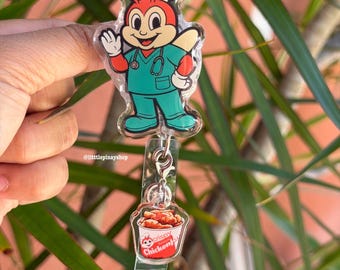May include: A clear badge reel featuring a cartoon character in a green medical uniform with a stethoscope. The character is wearing a white hat with a red cross. Below is a small bucket of crispy chicken. The text "Crispy Chickenjoy" is visible.