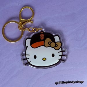 May include: An acrylic keychain featuring a white cat character wearing a black and orange baseball cap with an orange SF logo and a tan bow. The keychain includes a gold-toned metal ring and chain. The text @littlepinayshop is visible.