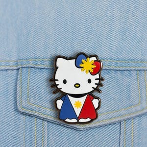 K&ouml;nnte beinhalten: Ein Hello Kitty Emaille-Anstecknadel mit wei&szlig;em Gesicht, schwarzen Schnurrhaaren und einer Schleife mit blauen, roten und gelben Akzenten. Die Anstecknadel hat ein Design mit blauen und roten Abschnitten und einem gelben Stern in der Mitte. Die Anstecknadel befindet sich auf einer hellblauen Jeansjacke.