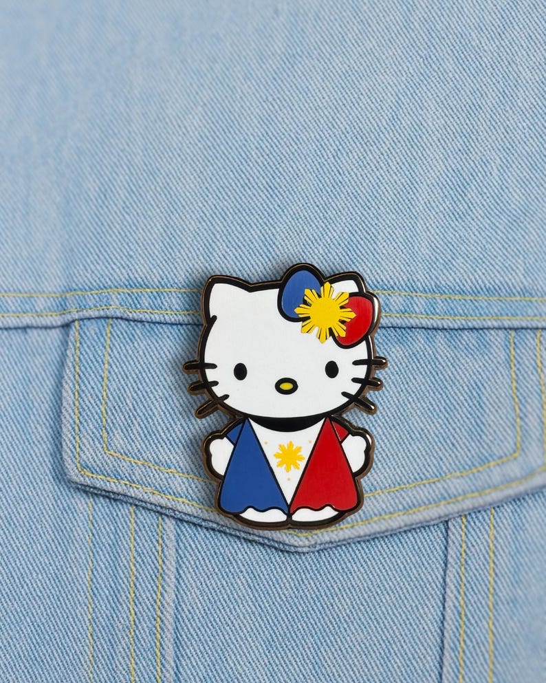 K&ouml;nnte beinhalten: Ein Hello Kitty Emaille-Anstecknadel mit wei&szlig;em Gesicht, schwarzen Schnurrhaaren und einer Schleife mit blauen, roten und gelben Akzenten. Die Anstecknadel hat ein Design mit blauen und roten Abschnitten und einem gelben Stern in der Mitte. Die Anstecknadel befindet sich auf einer hellblauen Jeansjacke.