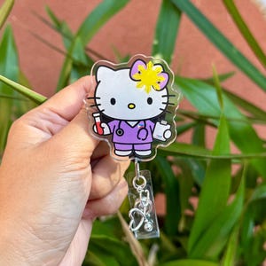 May include: A clear acrylic badge reel featuring a Hello Kitty design dressed as a nurse. The character wears a purple uniform and holds medical tools. The badge reel has a silver stethoscope charm and a clear plastic reel.