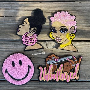 May include: Four embroidered patches on a wooden surface. The patches include two portraits of women, one with the text "Blessed & Black" on her earring, and the other with the text "Love" on her earring. There is also a pink smiley face and a patch with the text "Unbothered".