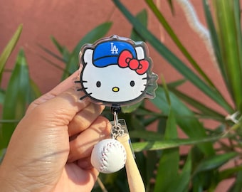 May include: A Hello Kitty badge reel featuring Hello Kitty wearing a blue baseball cap with the letters "LA" and a red bow. A small baseball and bat charm dangle below. The background is blurred with green plants.