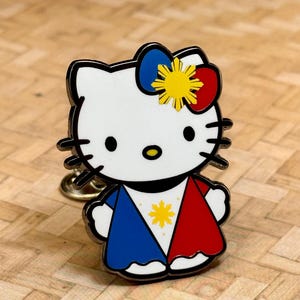 K&ouml;nnte beinhalten: Ein wei&szlig;er Emaille-Pin mit einer Cartoon-Katze mit schwarzem Umriss. Die Katze tr&auml;gt ein blaues und rotes Outfit mit einem gelben Stern und eine Schleife mit einem gelben Stern. Der Pin hat eine goldfarbene R&uuml;ckseite.