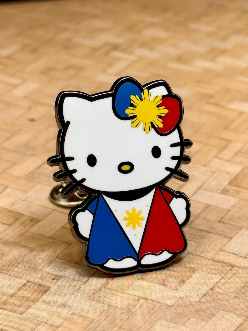 K&ouml;nnte beinhalten: Ein wei&szlig;er Emaille-Pin mit einer Cartoon-Katze mit schwarzem Umriss. Die Katze tr&auml;gt ein blaues und rotes Outfit mit einem gelben Stern und eine Schleife mit einem gelben Stern. Der Pin hat eine goldfarbene R&uuml;ckseite.