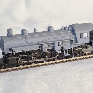 Puede incluir: Una maqueta gris de locomotora de vapor con detalles, incluyendo una chimenea, cabina y ténder. El tren modelo está sobre una sección de vía férrea en miniatura, mostrando su intrincado diseño. La locomotora es un modelo a escala.