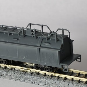 Puede incluir: Un vagón cisterna de tren modelo gris con detalles, incluyendo una escalera y barandillas en la parte superior. El vagón está sobre una sección de vía de tren modelo, mostrando las ruedas y el tren de rodaje.