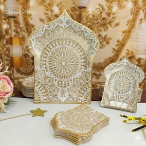 Può includere: Piatti e tovaglioli di carta decorativi bianchi e oro con un intricato design a mandala. I piatti hanno la forma di un arco. Il testo "Damai Party" è visibile nell'angolo in alto a sinistra.