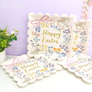 May include: Scalloped-edge paper plates with a floral and bunny design. The plates feature a pink bow, the words "Happy Easter" in gold, and illustrations of flowers, eggs, and rabbits. Perfect for Easter celebrations.