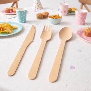 Op de afbeelding: Een feestelijke verjaardagstafel met houten bestek: een mes, vork en lepel. De tafel is gedekt met borden, bekers, snacks en cupcakes. Een kleurrijke "Happy Birthday" banner hangt op de achtergrond.