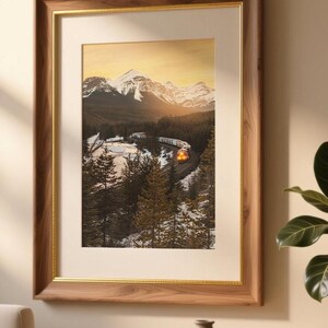 May include: Framed landscape art print featuring a train winding through a snowy mountain valley. The artwork is encased in a wooden frame with a gold inner border. The scene depicts snow-capped mountains, evergreen trees, and a warm sunset.