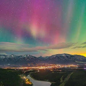 Pode incluir: Uma paisagem noturna com uma aurora boreal vibrante com faixas de luz roxa, rosa e verde sobre uma cordilheira. Abaixo dos picos, uma cidade brilha com luzes amarelas quentes, situada perto de um rio escuro que serpenteia por uma floresta.