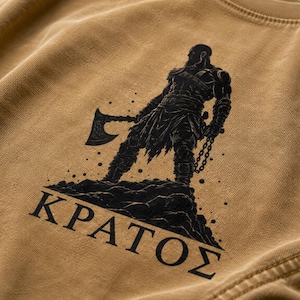 May include: A tan-colored t-shirt featuring a black graphic print of a muscular warrior figure standing on a rocky mound. The figure holds an axe in one hand and a chain in the other. Below the figure, the Greek text "&Kappa;&Rho;&Alpha;&Tau;&Omicron;&Sigma;" is printed in black.