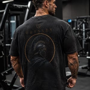 May include: A black short-sleeve t-shirt with a faded wash finish. The back features a graphic of a Spartan helmet inside a gold circle, with the word ODYSSEY printed in gold lettering above the circle and a small logo icon at the top.