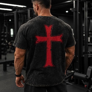 Könnte beinhalten: Ein kurzärmeliges T-Shirt in schwarzer Acid-Wash-Optik mit einem großen, abgenutzten roten Kreuz-Motiv auf der Rückseite. Das Kleidungsstück hat einen Rundhalsausschnitt und eine Standardpassform, wobei der Stoff eine texturierte Optik aufweist.