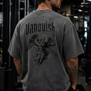 Puede incluir: Una camiseta gris de gran tamaño con la palabra "Vanquish" en una fuente gótica en la espalda. Debajo del texto hay una ilustración detallada de un ángel con grandes alas. La camiseta la lleva una persona en un gimnasio.