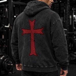 May include: A charcoal grey acid-wash hooded sweatshirt featuring a large, distressed red cross graphic printed on the back. The long-sleeved pullover has a hood, ribbed cuffs, and a ribbed hem, showcasing a worn-in texture and a relaxed fit.