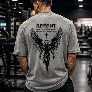 May include: A grey short-sleeve t-shirt featuring a black graphic on the back. The design depicts a winged figure with a halo and a cross above it, accompanied by the text "REPENT for the Kingdom of Heaven has come near" in black lettering.