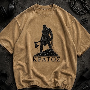 May include: A tan, acid-wash t-shirt featuring a black graphic print of a warrior holding an axe and chains, standing on a rocky mound. Below the figure, the Greek text "&Kappa;&Rho;&Alpha;&Tau;&Omicron;&Sigma;" is printed in bold, black, capital letters on the center chest.