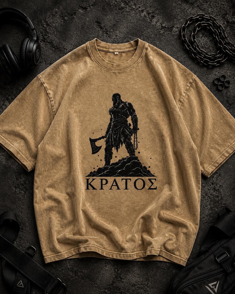 May include: A tan, acid-wash t-shirt featuring a black graphic print of a warrior holding an axe and chains, standing on a rocky mound. Below the figure, the Greek text "&Kappa;&Rho;&Alpha;&Tau;&Omicron;&Sigma;" is printed in bold, black, capital letters on the center chest.
