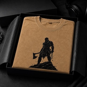 May include: A tan crew neck t-shirt featuring a black graphic print of a warrior figure standing on a rocky mound, holding a large axe in one hand and a chain in the other, presented neatly folded inside a black gift box with black tissue paper.