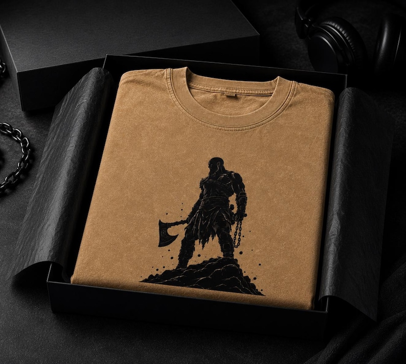 May include: A tan crew neck t-shirt featuring a black graphic print of a warrior figure standing on a rocky mound, holding a large axe in one hand and a chain in the other, presented neatly folded inside a black gift box with black tissue paper.