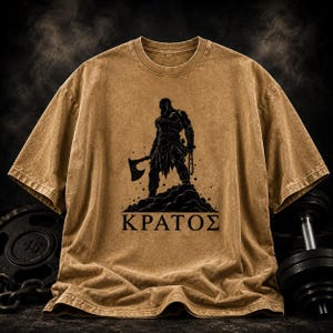 May include: A tan, mineral-washed t-shirt featuring a black graphic print of a muscular warrior holding an axe and standing on a rocky mound. Below the figure, the Greek text "&Kappa;&Rho;&Alpha;&Tau;&Omicron;&Sigma;" is printed in a bold, black, sans-serif typeface.