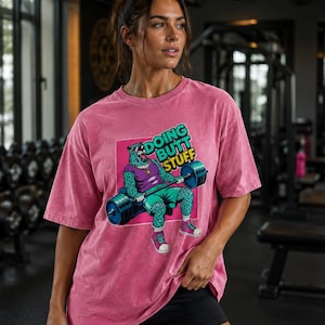 May include: A pink short-sleeve t-shirt featuring a graphic of a teal leopard wearing a purple tank top and sunglasses, lifting a blue barbell. The text "DOING BUTT STUFF" is printed in green and yellow above the leopard on a pink square background.