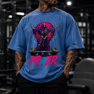 Könnte beinhalten: Ein blaues Oversized-T-Shirt mit einer Grafik eines Skeletts in einer violetten Robe mit erhobenen Armen, das hinter einer Langhantel steht. Das Design zeigt einen pinken Kreis, Blitze und den Schriftzug PR OR ER in fetten pinken Buchstaben.