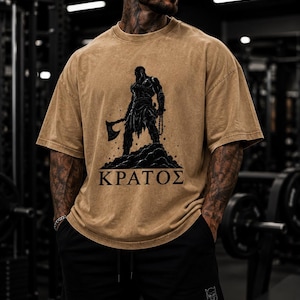 May include: A tan, acid-wash style t-shirt featuring a black graphic illustration of a warrior figure holding an axe, standing on a rocky mound, with the Greek text "&Kappa;&Rho;&Alpha;&Tau;&Omicron;&Sigma;" printed in black capital letters below the graphic design.