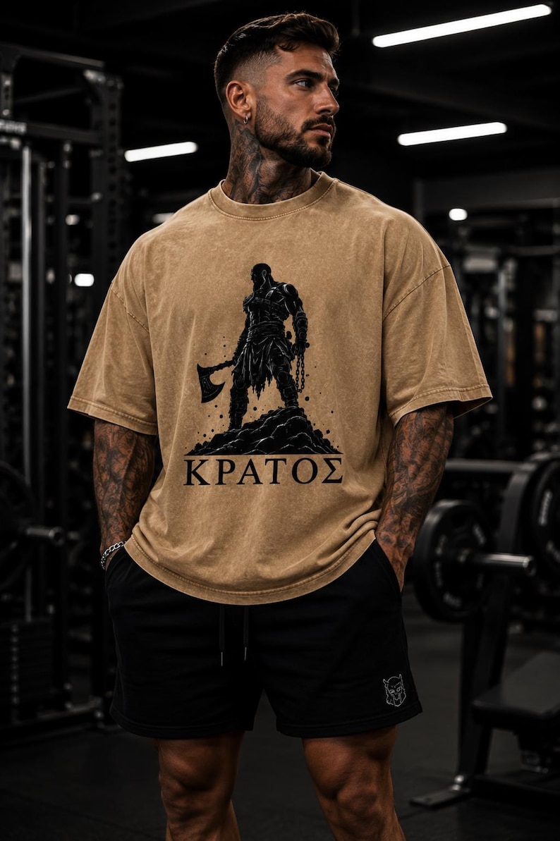 May include: A tan, acid-wash style t-shirt featuring a black graphic illustration of a warrior figure holding an axe, standing on a rocky mound, with the Greek text "&Kappa;&Rho;&Alpha;&Tau;&Omicron;&Sigma;" printed in black capital letters below the graphic design.