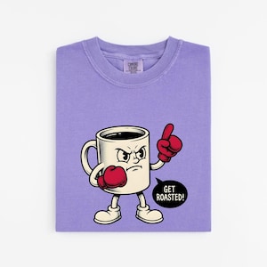 May include: A light purple t-shirt featuring a graphic of an angry white coffee mug character wearing red boxing gloves. The mug has arms and legs, is pointing a finger, and has a black speech bubble with the white text GET ROASTED!