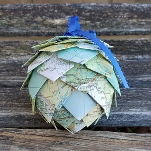 May include: A blue ribbon ornament made from folded paper maps of Finland. The ornament features the names of cities and towns in Finland, including Helsinki, Tampere, and Turku.