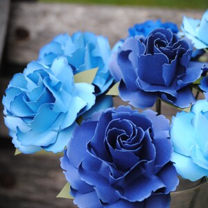 May include: A bouquet of blue and light blue paper flowers in a clear glass vase. The flowers are arranged in a cluster and have a realistic appearance.