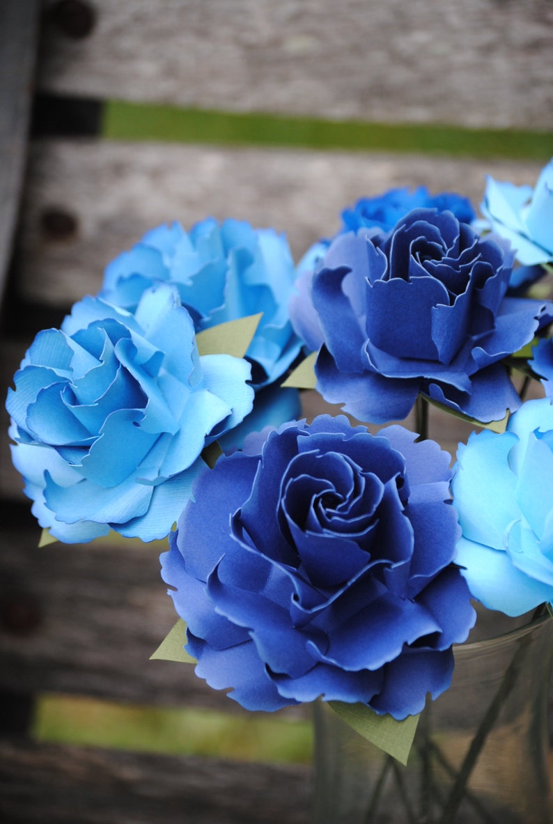 May include: A bouquet of blue and light blue paper flowers in a clear glass vase. The flowers are arranged in a cluster and have a realistic appearance.
