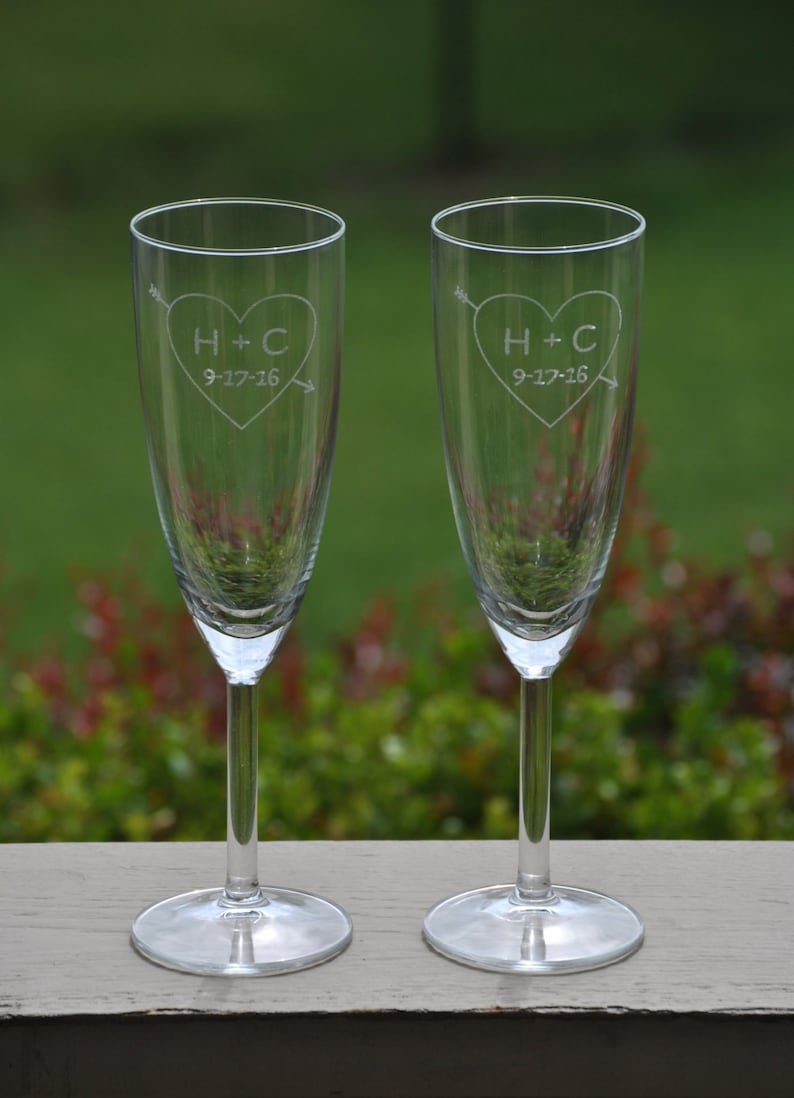 May include: Two clear glass champagne flutes with etched hearts and the initials H+C and the date 9-17-16. The flutes are on a wooden surface.