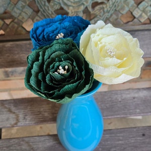 May include: Three paper flowers in a blue vase. The flowers are teal, green, and cream. The flowers are arranged in a bouquet.