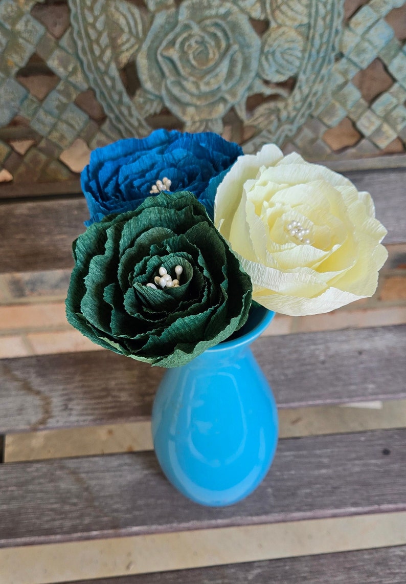 May include: Three paper flowers in a blue vase. The flowers are teal, green, and cream. The flowers are arranged in a bouquet.