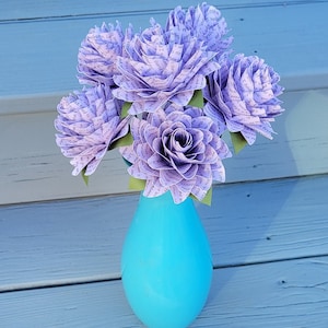 May include: A bouquet of six lavender paper flowers with text printed on the petals, arranged in a turquoise vase. The vase is sitting on a light gray wooden surface.