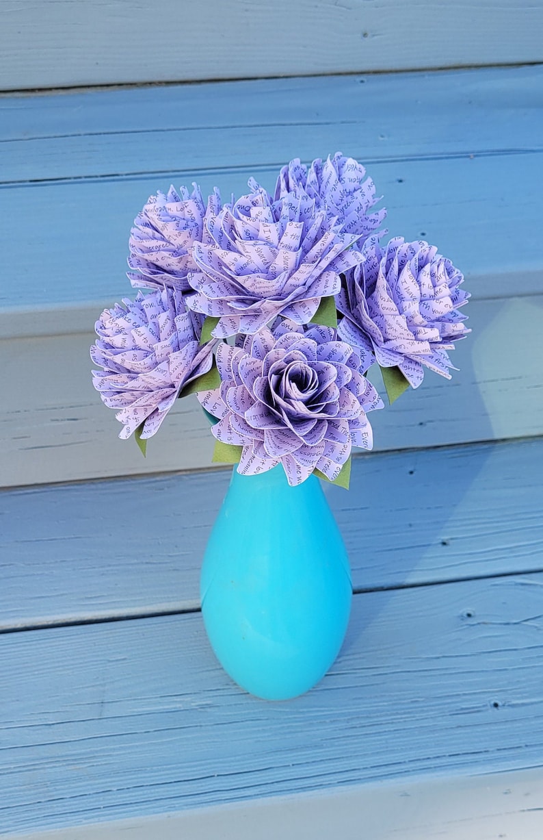 May include: A bouquet of six lavender paper flowers with text printed on the petals, arranged in a turquoise vase. The vase is sitting on a light gray wooden surface.
