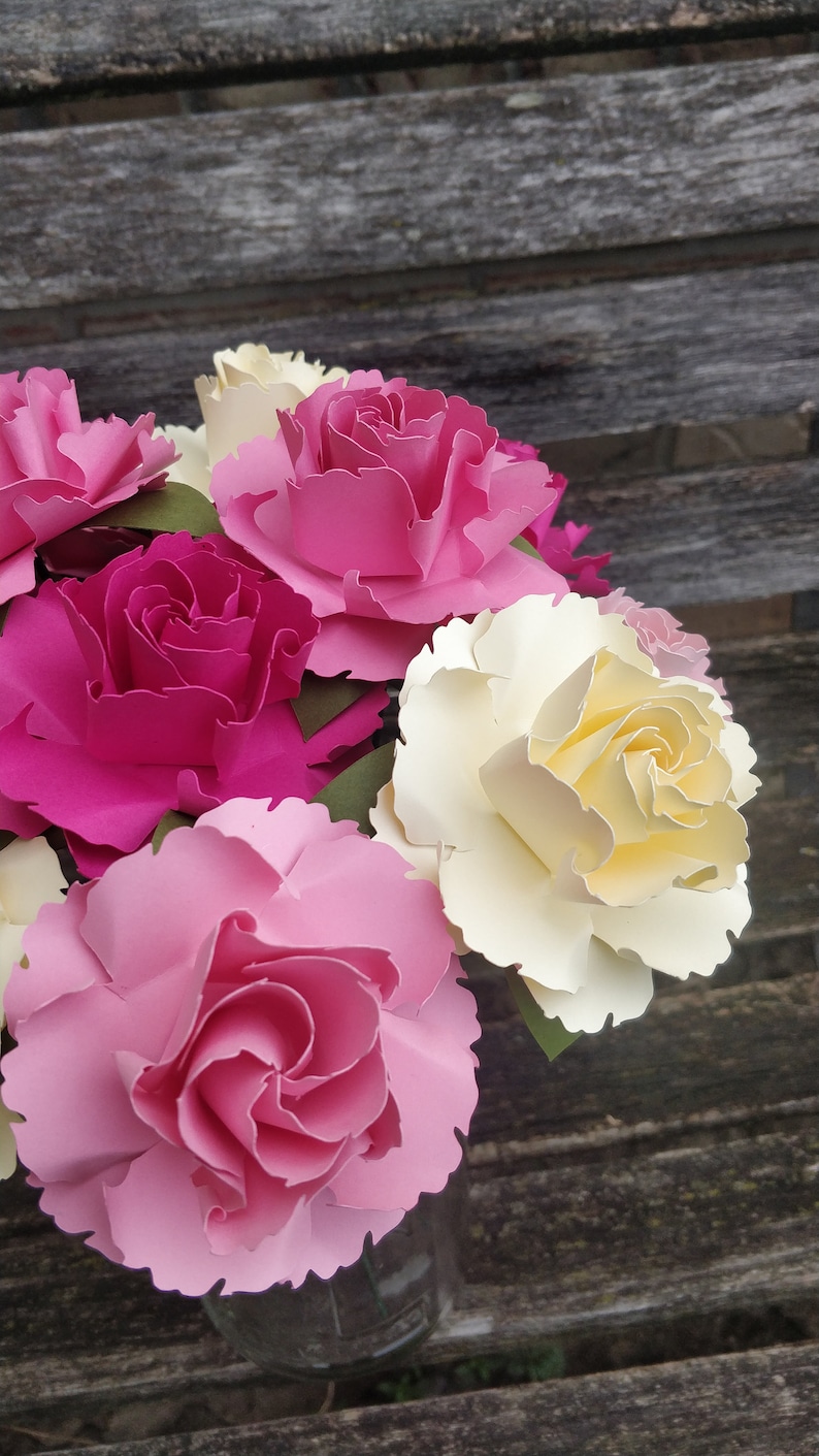 May include: A bouquet of paper flowers in a glass vase. The flowers are pink, white, and yellow. The flowers are arranged in a vase on a wooden surface.