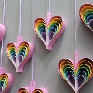 K&ouml;nnte beinhalten: Eine Wand, die mit mehreren Regenbogen-Papierherzen geschm&uuml;ckt ist, die an Schn&uuml;ren h&auml;ngen. Jedes Herz besteht aus &uuml;bereinanderliegenden Papier in den Farben Rot, Orange, Gelb, Gr&uuml;n, Blau, Indigo und Violett.