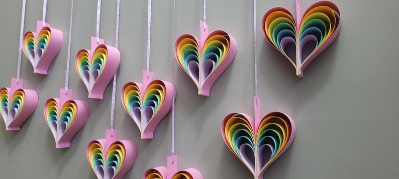 K&ouml;nnte beinhalten: Eine Wand, die mit mehreren Regenbogen-Papierherzen geschm&uuml;ckt ist, die an Schn&uuml;ren h&auml;ngen. Jedes Herz besteht aus &uuml;bereinanderliegenden Papier in den Farben Rot, Orange, Gelb, Gr&uuml;n, Blau, Indigo und Violett.